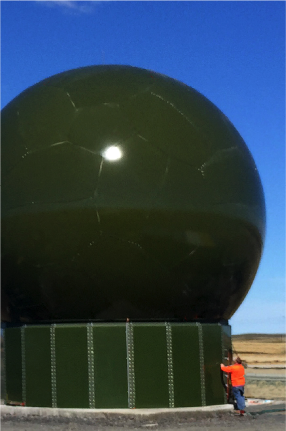 A person in an orange shirt stands next to a large, green, dome-shaped radar structure under a clear blue sky, showcasing the impressive specifications and size difference between the two.