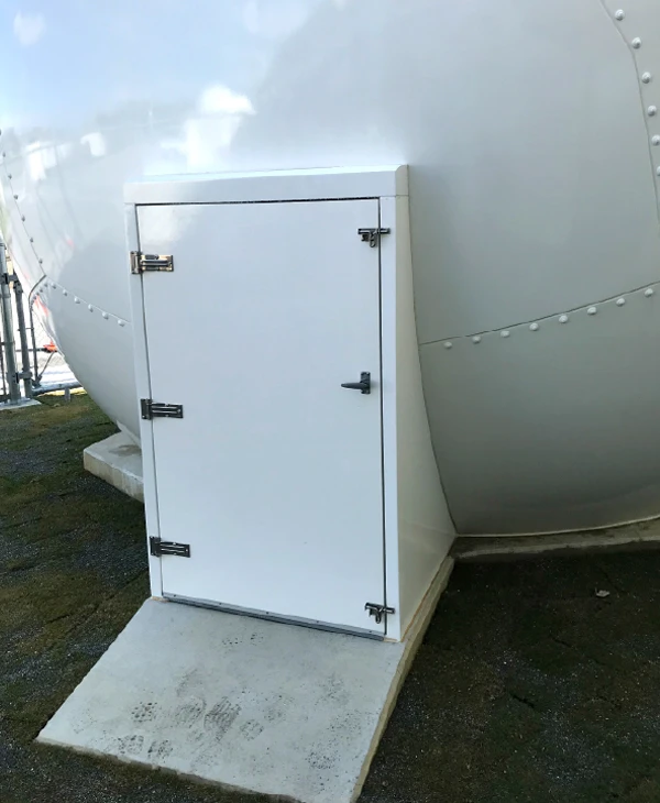 A small white door with metal hinges sits at the base of a large, smooth radome on a concrete platform outdoors.